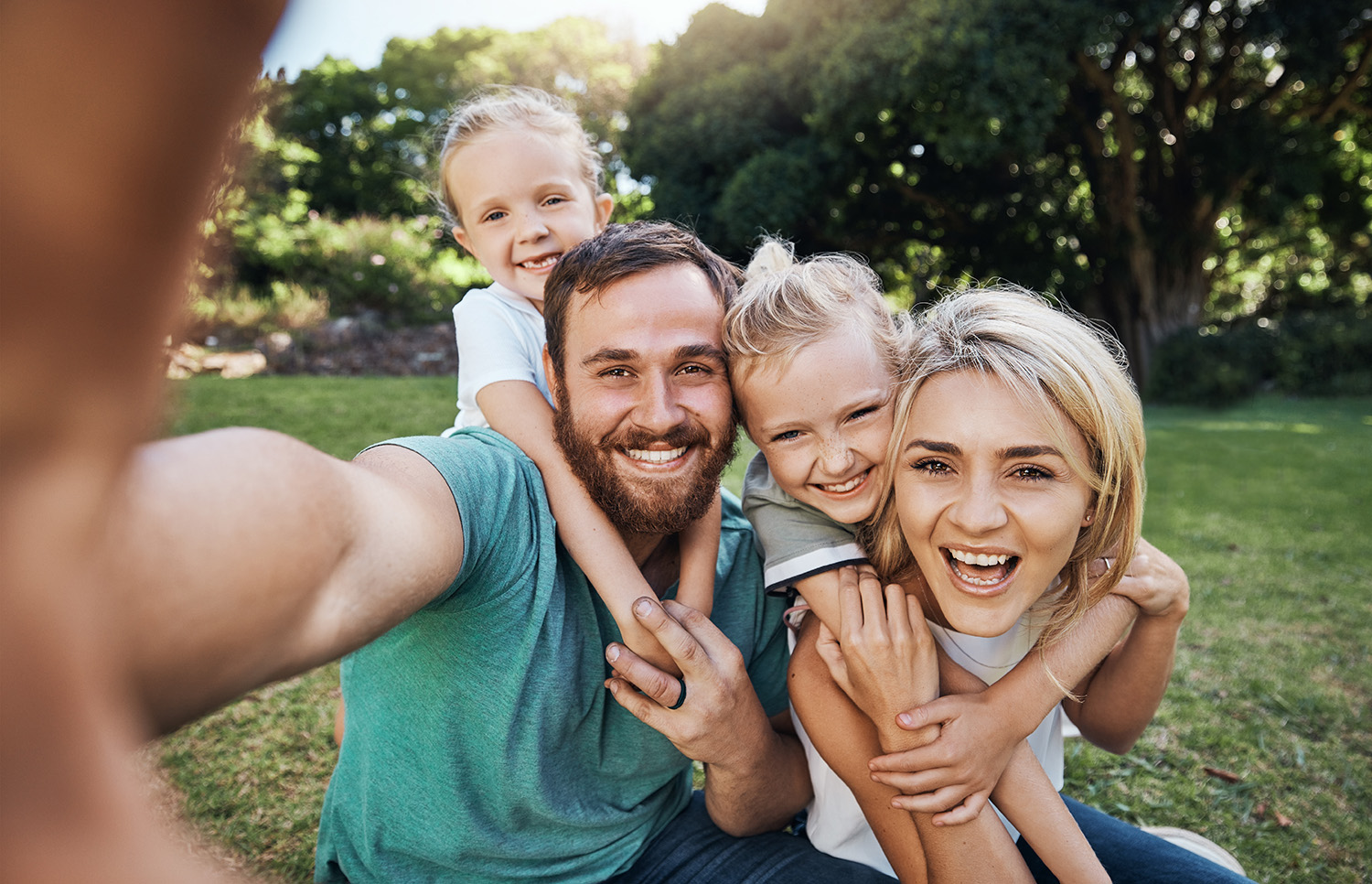 Family with young children take a selfie in a park with trees and grass.
